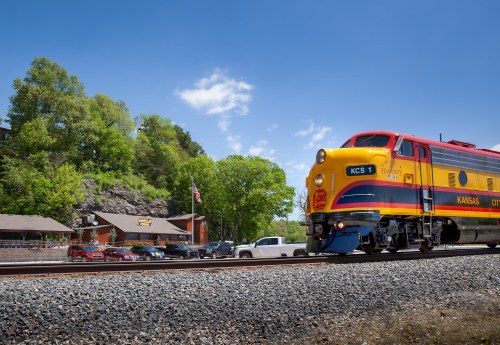 A colorful Kansas City Southern train passes by a rustic station surrounded by green trees and parked cars.