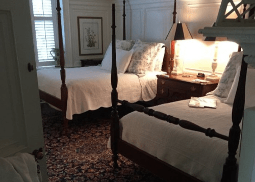 A cozy bedroom featuring two four-poster beds, a nightstand, and soft lighting, with a patterned rug on the floor.