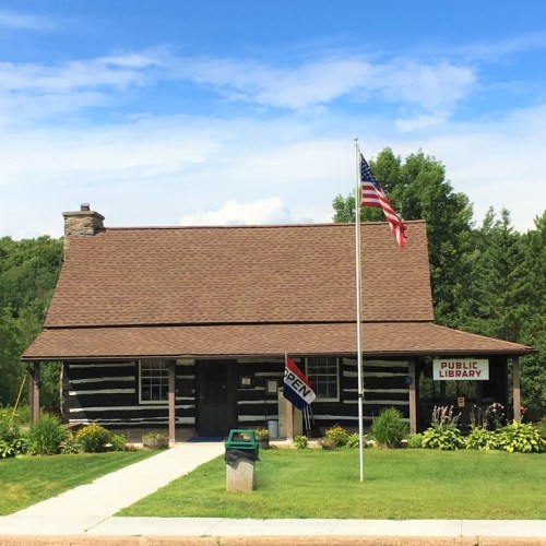 A rustic log cabin library with a flag, surrounded by greenery and a clear blue sky. A sign indicates it's open.