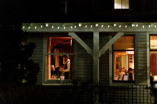 A cozy dinner scene viewed through a window, with people gathered around a table, illuminated by warm lights.