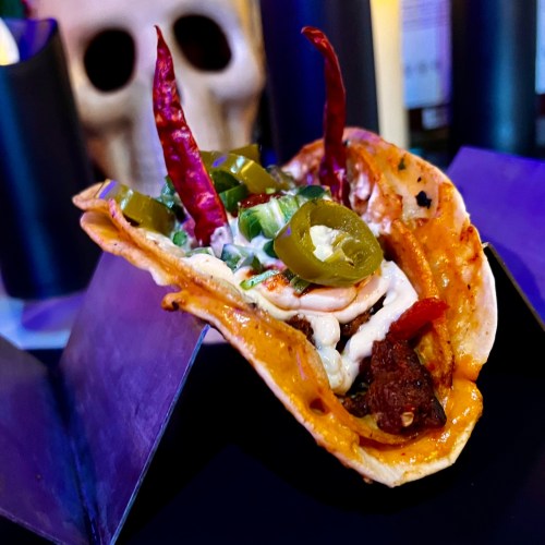 A close-up of a taco filled with meat, jalapeños, and topped with red chilies, served on a dark background.