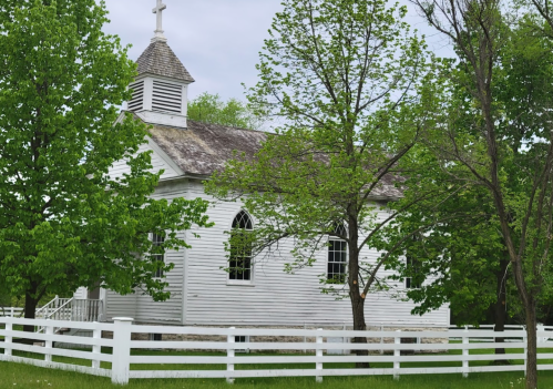 A white church surrounded by green trees and a white fence on a cloudy day.
