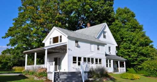 A white two-story house with a porch, surrounded by green trees and a clear blue sky.