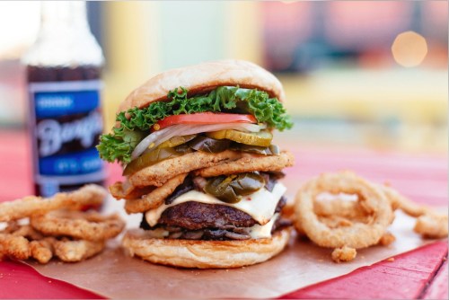 A towering burger with lettuce, tomato, pickles, and onion rings, served with a bottle of soda and onion rings on the side.