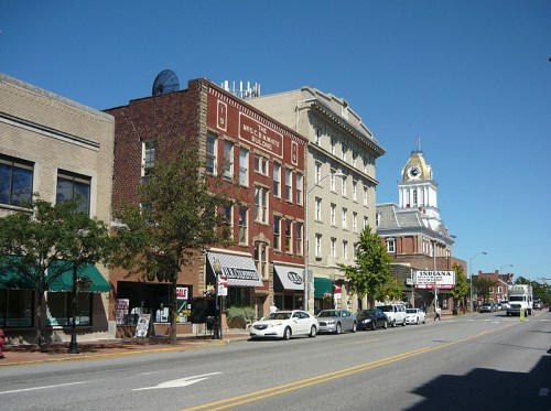A sunny street scene featuring historic buildings, shops, and a clock tower in the background.
