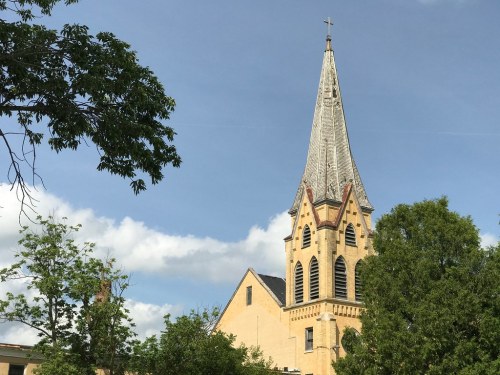 A tall church steeple with a cross, surrounded by trees and a blue sky with clouds.