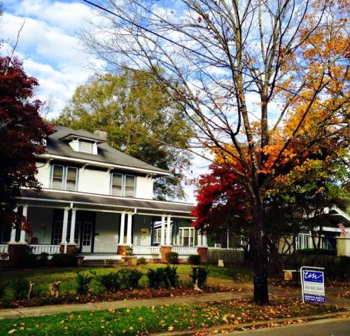 A charming two-story house with a porch, surrounded by colorful autumn trees and a real estate sign in the front yard.