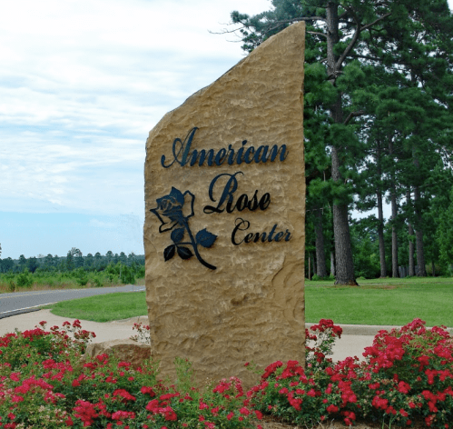 Sign for the American Rose Center, featuring a rose design, surrounded by blooming red flowers and greenery.