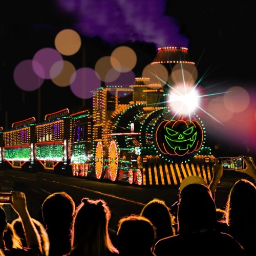 A brightly lit train with a pumpkin face, surrounded by colorful lights, as people watch and take photos.