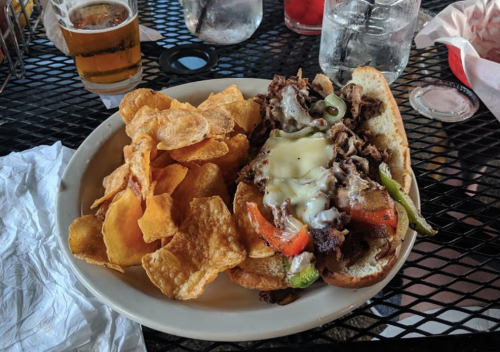 A plate with a loaded sandwich filled with beef, peppers, and cheese, accompanied by a side of crispy potato chips.
