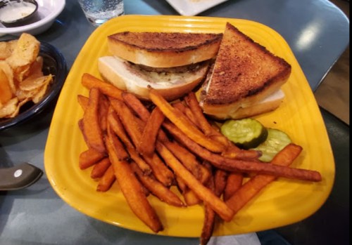 A yellow plate with two toasted sandwiches, sweet potato fries, and pickles on the side.