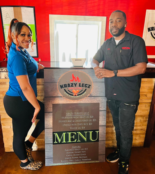 A woman and man stand beside a menu board at Krazy Legz, showcasing various food items and prices.