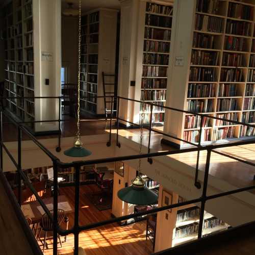 A spacious library with tall bookshelves, wooden floors, and green pendant lights, bathed in warm sunlight.