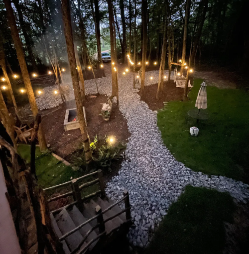 A serene outdoor space with a gravel path, fire pit, and string lights among trees, creating a cozy atmosphere.
