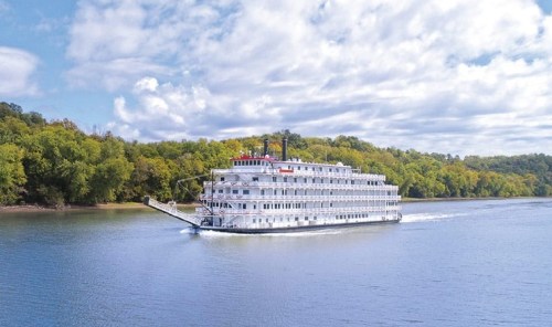 A large riverboat cruises along a calm river, surrounded by lush green trees and a partly cloudy sky.