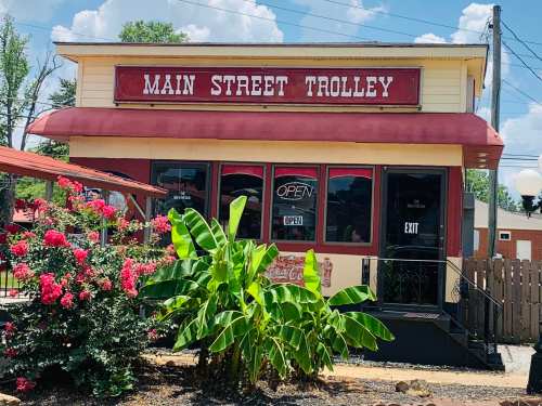 A quaint restaurant named "Main Street Trolley" with a red awning, surrounded by flowers and greenery.
