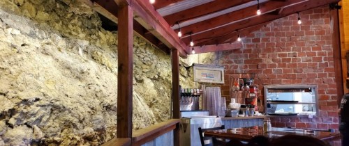 Interior of a rustic café with exposed brick and rock walls, wooden beams, and warm lighting.