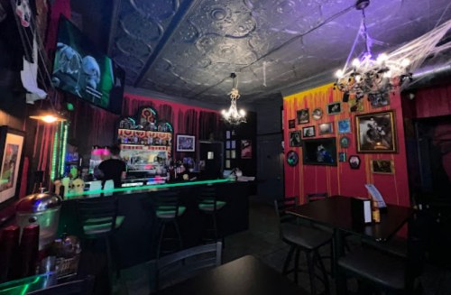 A dimly lit bar with colorful decor, chandeliers, and Halloween-themed artwork on the walls.