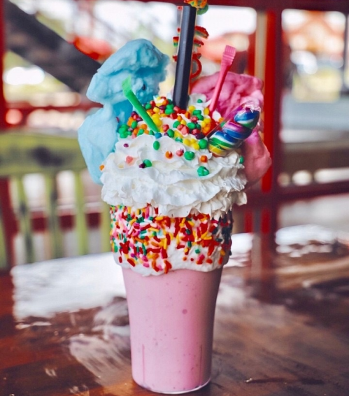 A colorful milkshake topped with whipped cream, cotton candy, sprinkles, and a candy swirl, served in a tall cup.