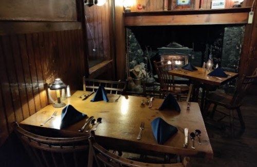 Cozy restaurant interior with wooden walls, tables set with blue napkins, and a fireplace in the background.