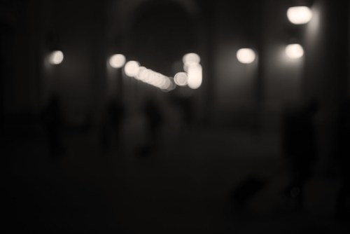 A blurred, dimly lit corridor with soft glowing lights and indistinct figures in the background.