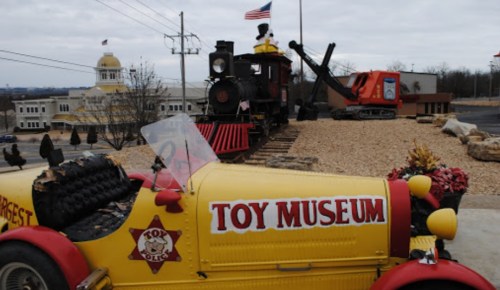 A colorful toy museum display featuring a vintage car and a train on tracks, with a building and flag in the background.