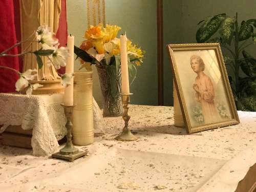 A table with candles, a vase of flowers, and a framed portrait of a young girl, set against a colorful backdrop.
