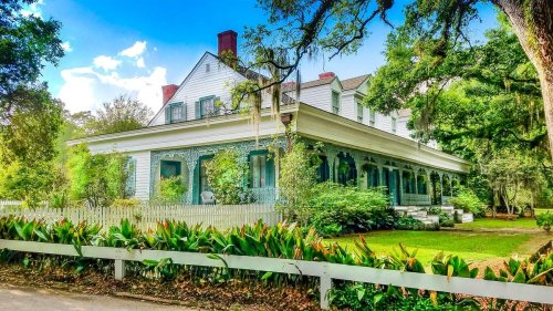 A charming two-story house with a wraparound porch, surrounded by lush greenery and trees under a bright blue sky.