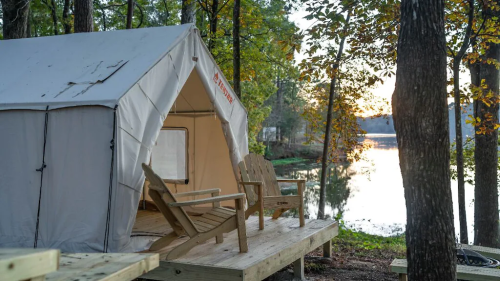A cozy tent on a wooden deck by a serene lake, surrounded by trees and autumn foliage at sunset.