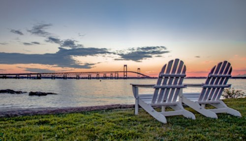 Two white Adirondack chairs facing a serene sunset over a bridge and calm water, with clouds in the sky.