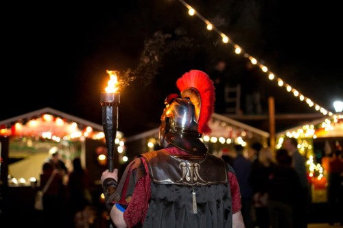 A person in a Roman soldier costume holds a torch, surrounded by festive lights and a crowd at night.