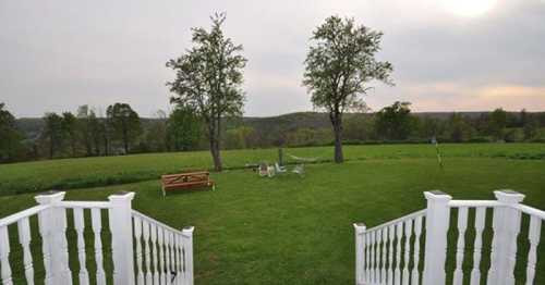 A view of a grassy field with two trees, a bench, and distant hills under a cloudy sky. Steps lead down from a porch.