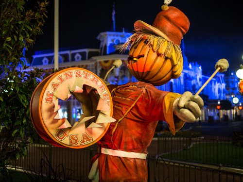 A whimsical scarecrow figure holding a drum, set against a nighttime backdrop of colorful lights.