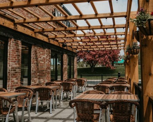 Outdoor dining area with wooden trellis, tables, and chairs, surrounded by brick walls and a blooming tree in the background.