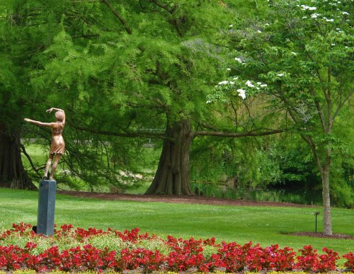 A bronze statue of a dancer stands amidst vibrant flowers and lush green trees by a serene pond.