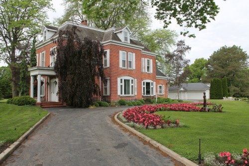 A large brick house with white trim, surrounded by green lawns and colorful flower beds, on a cloudy day.