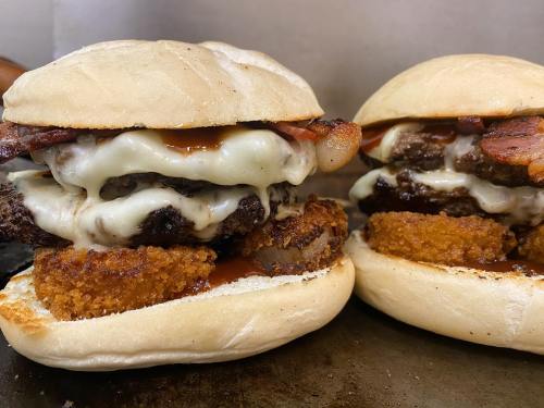 Two stacked burgers with melted cheese, bacon, and crispy onion rings on soft buns, drizzled with barbecue sauce.