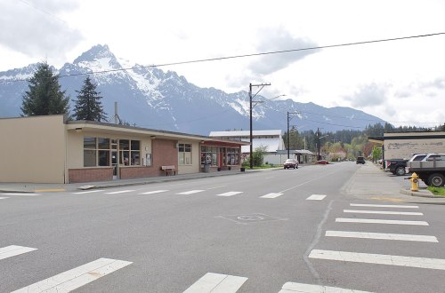 A quiet street scene with buildings and mountains in the background, featuring snow-capped peaks under a cloudy sky.