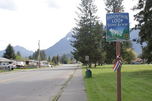 A scenic road sign for Mountain Loop, with mountains and trees in the background, along a quiet street.