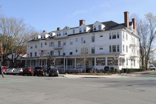 A large white hotel building with multiple stories, surrounded by trees and parked cars on a sunny day.
