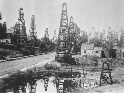 Historic oil derricks line a dirt road, with a small pond reflecting the structures in a black-and-white photograph.
