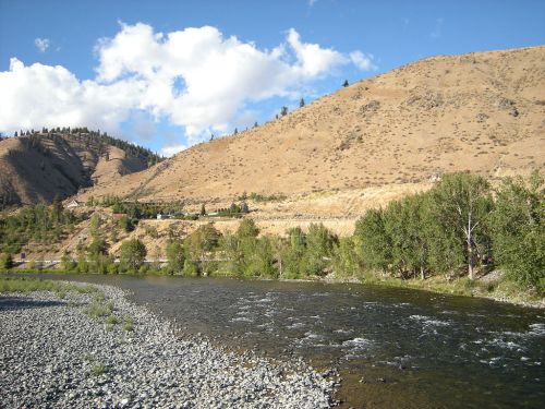 A serene river flows through a rocky landscape, bordered by dry hills and scattered trees under a partly cloudy sky.