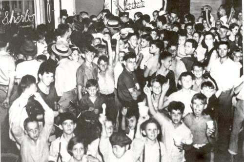 A crowded black-and-white photo of a large group of children and teens, some holding ice cream, in a lively setting.