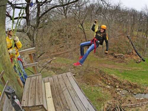 A person in a helmet zip lines from a platform while another person prepares on the ground, surrounded by trees.