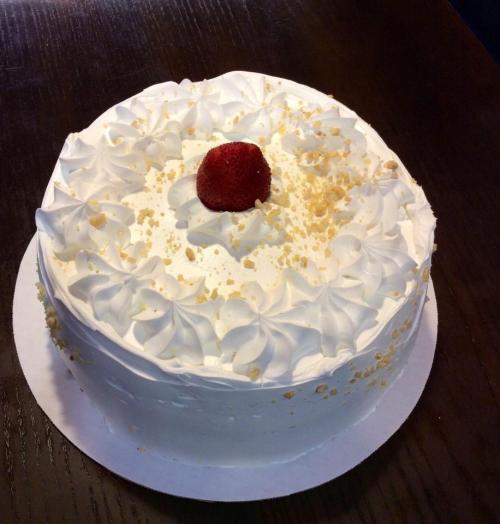 A round white cake topped with whipped cream, a strawberry, and golden crumbs on a wooden table.