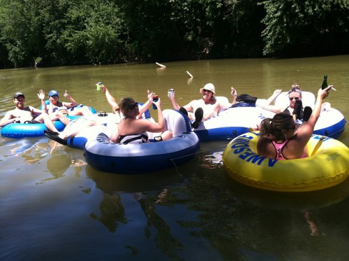 A group of people relaxes on inflatable tubes in a river, enjoying drinks and waving at the camera.