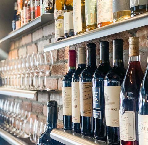 A shelf displaying various wine bottles and glasses against a brick wall.
