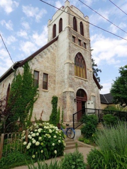 A historic brick church with a tall tower, surrounded by greenery and flowers, under a partly cloudy sky.