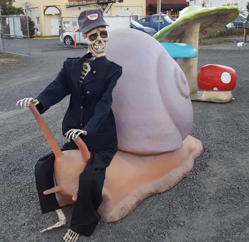 A skeleton in a taxi driver outfit sits on a large snail sculpture in a gravel area.