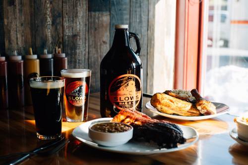 A table with BBQ food, two beers, and a large growler from Slows BBQ, with a rustic wooden backdrop.